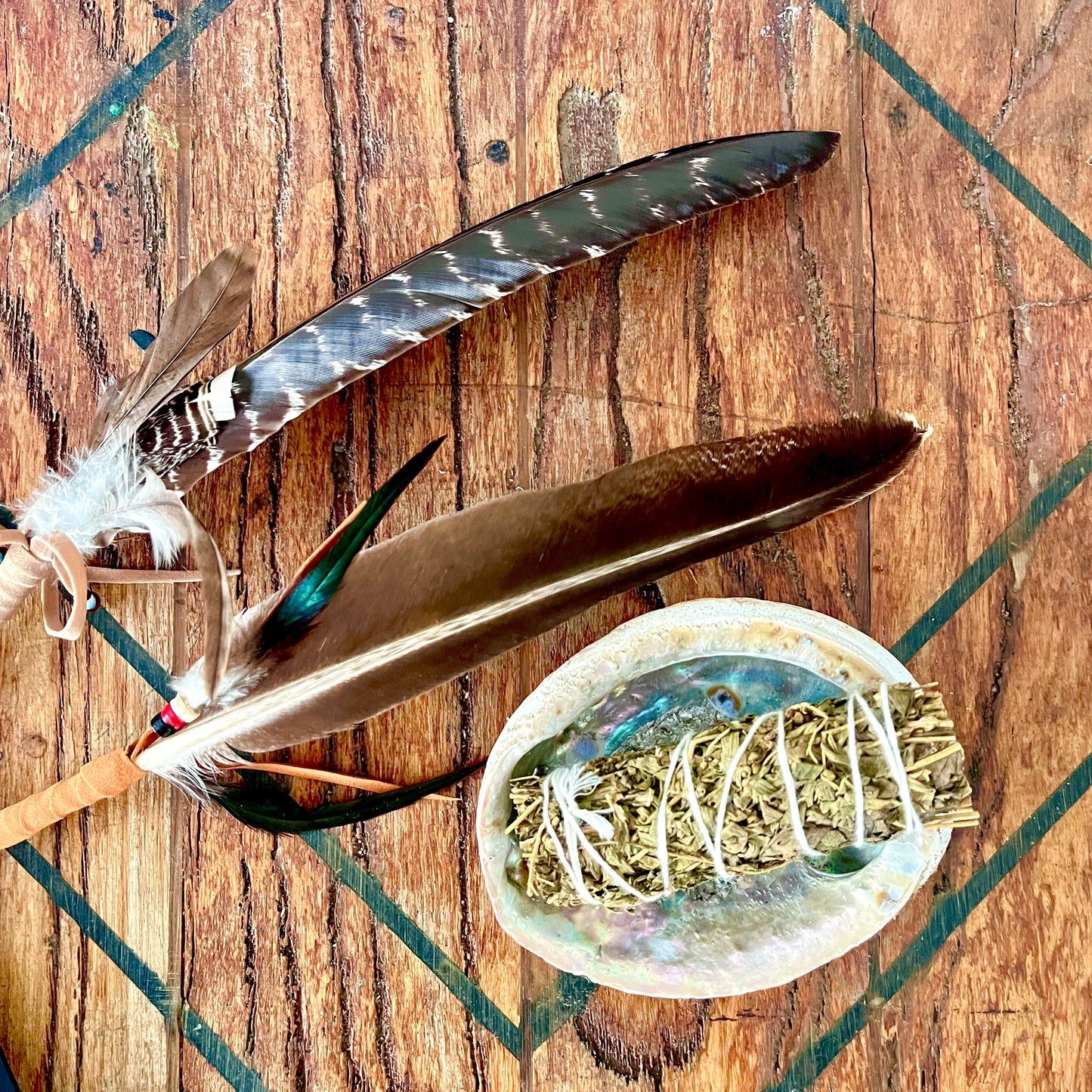 A natural leather-wrapped bundle with two large brown and gray feathers, a small turquoise feather accent, and a white sage smudge holder inside an iridescent shell, arranged on a rustic wooden surface.