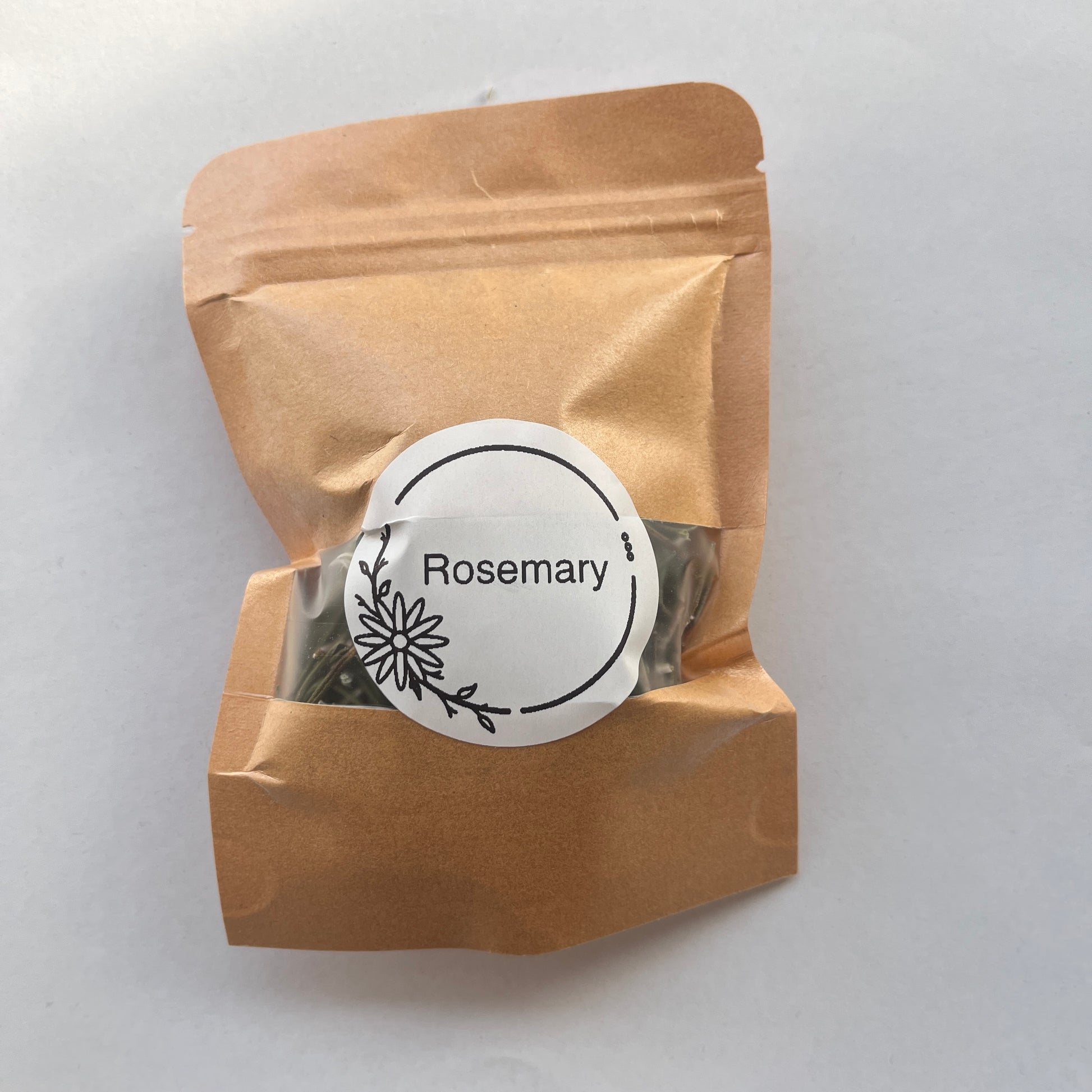 A small kraft paper resealable pouch with a clear window showing dried rosemary leaves inside, topped with a round white label that reads 'Rosemary' with a simple floral line drawing.