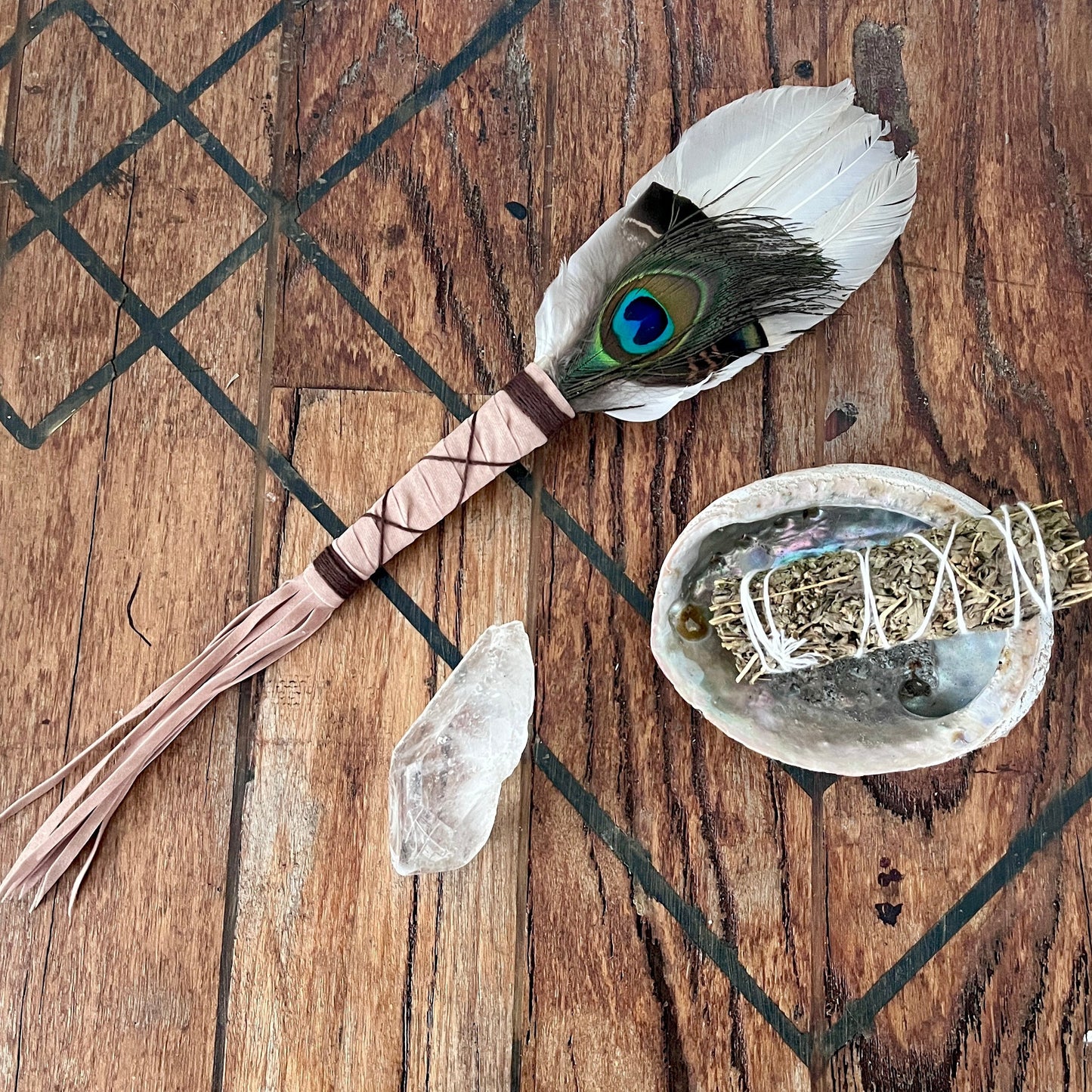 A handmade feather wand with a peacock feather and wrapped handle lies on a rustic wooden surface next to a white sage smudge bundle in an abalone shell, a clear crystal, and a black geometric design painted on the wood.