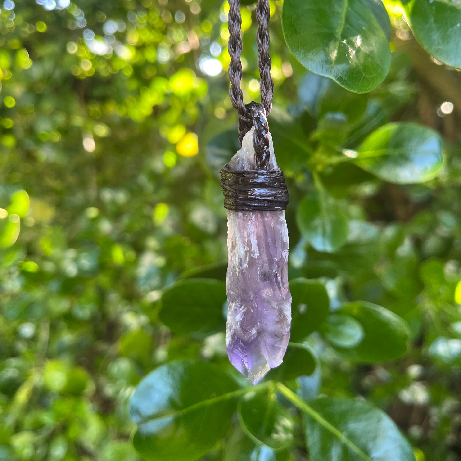A raw amethyst crystal pendant necklace hanging from a dark brown cord, set against a blurred background of green leaves.