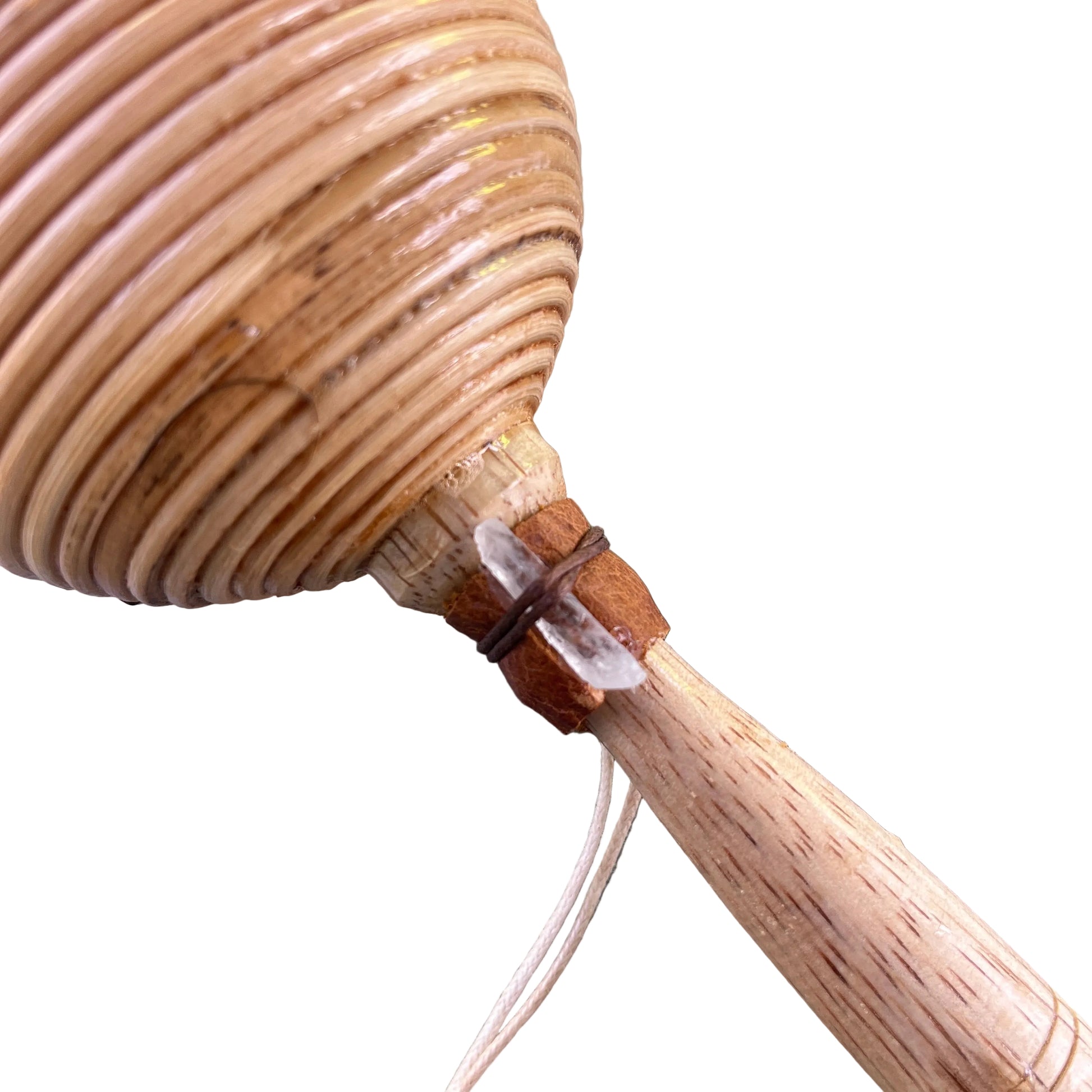 A close-up of a handmade wooden shamanic rattle shaker, a light wood handle, and a small clear crystal wrapped in brown thread, resting on a gray textured surface.