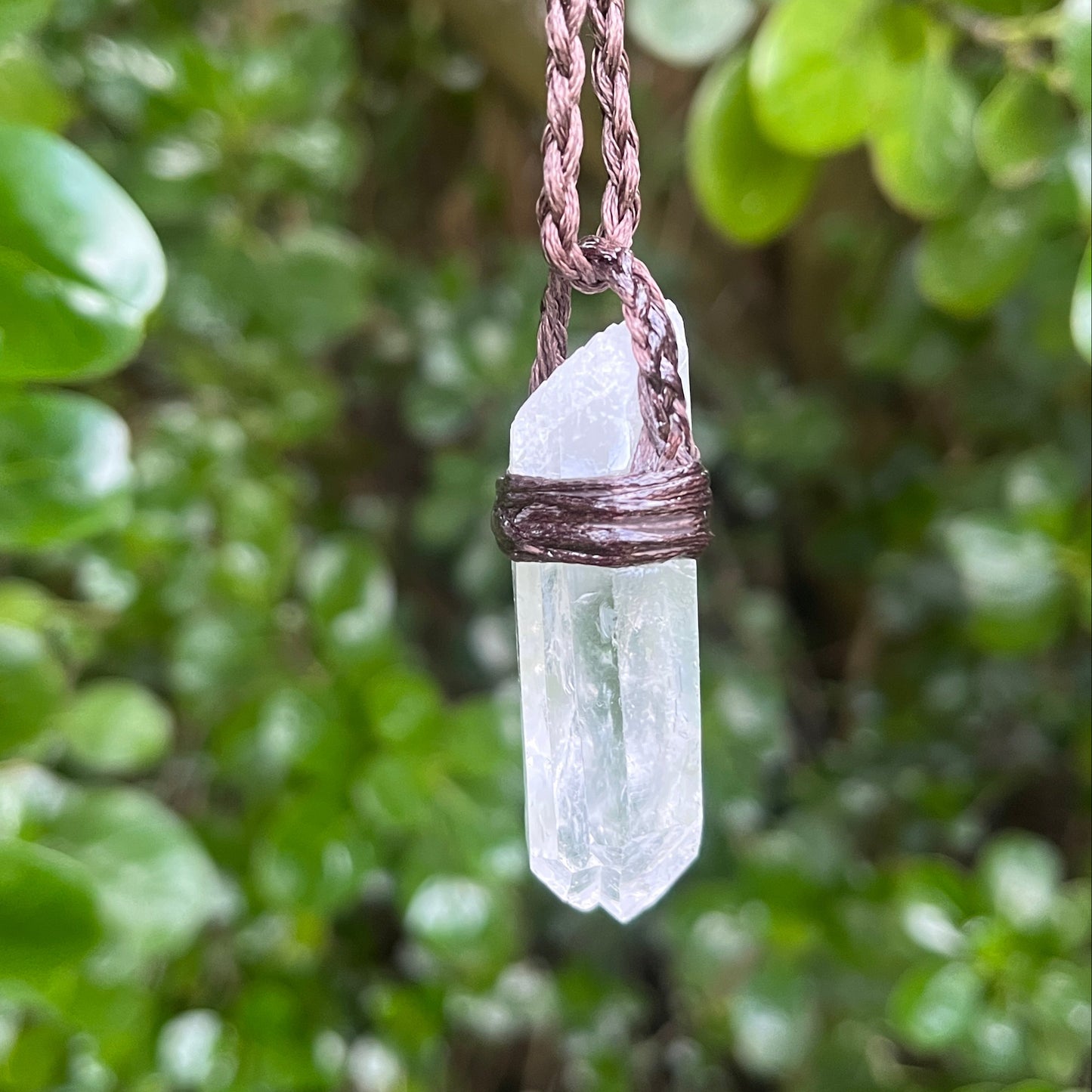 A clear quartz crystal pendant necklace with a brown braided cord and wrapped metal cap, hanging against a blurred background of green leaves.