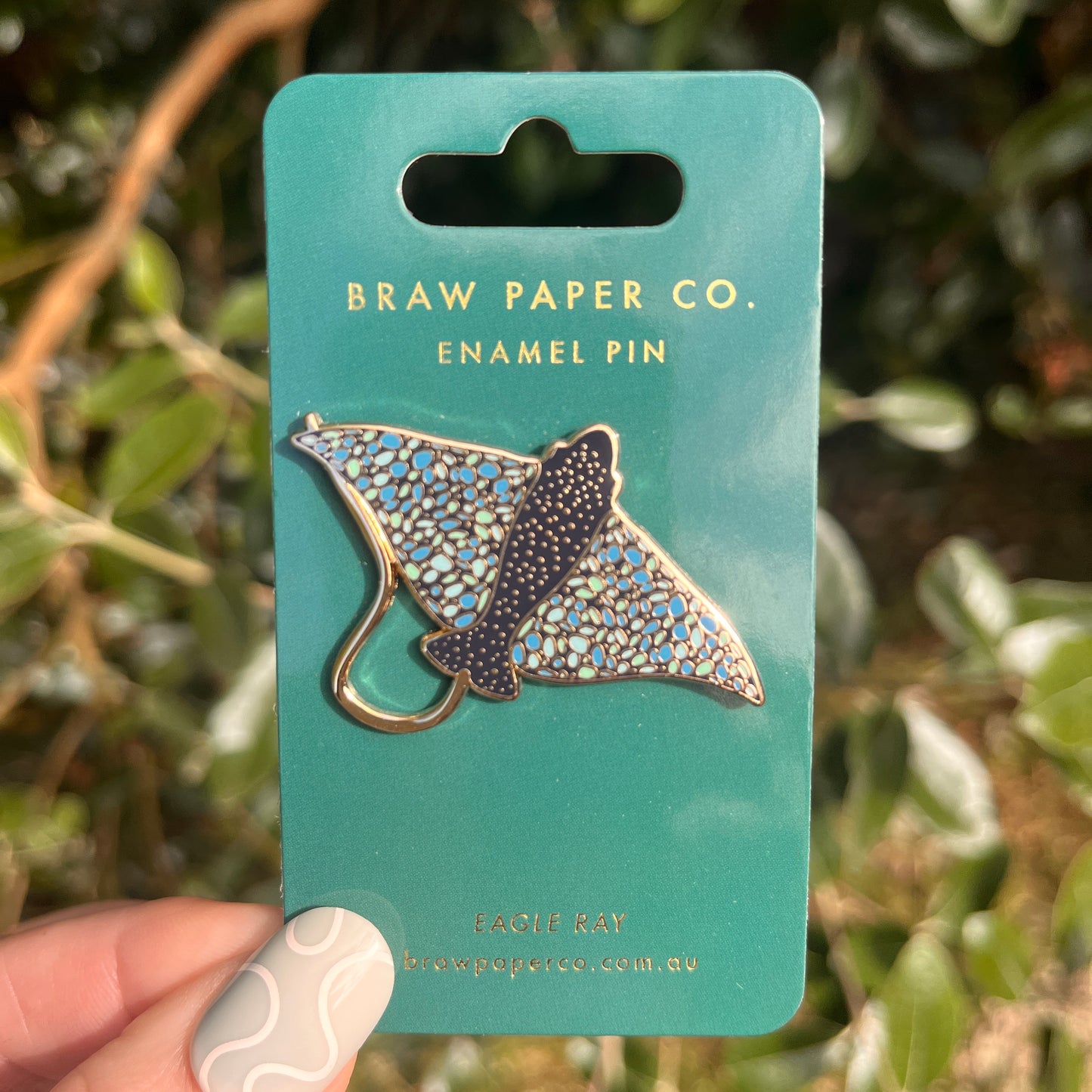 Enamel pin of an eagle ray on a Braw Paper Co. card with a blurred green leafy background