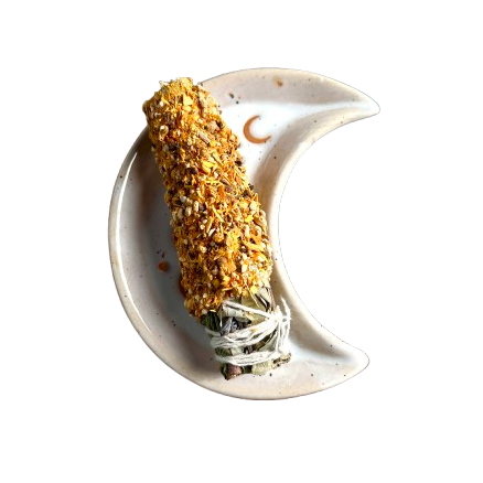 A dried herb smudge bundle wrapped in white string, resting on a ceramic crescent moon dish against a solid white background.