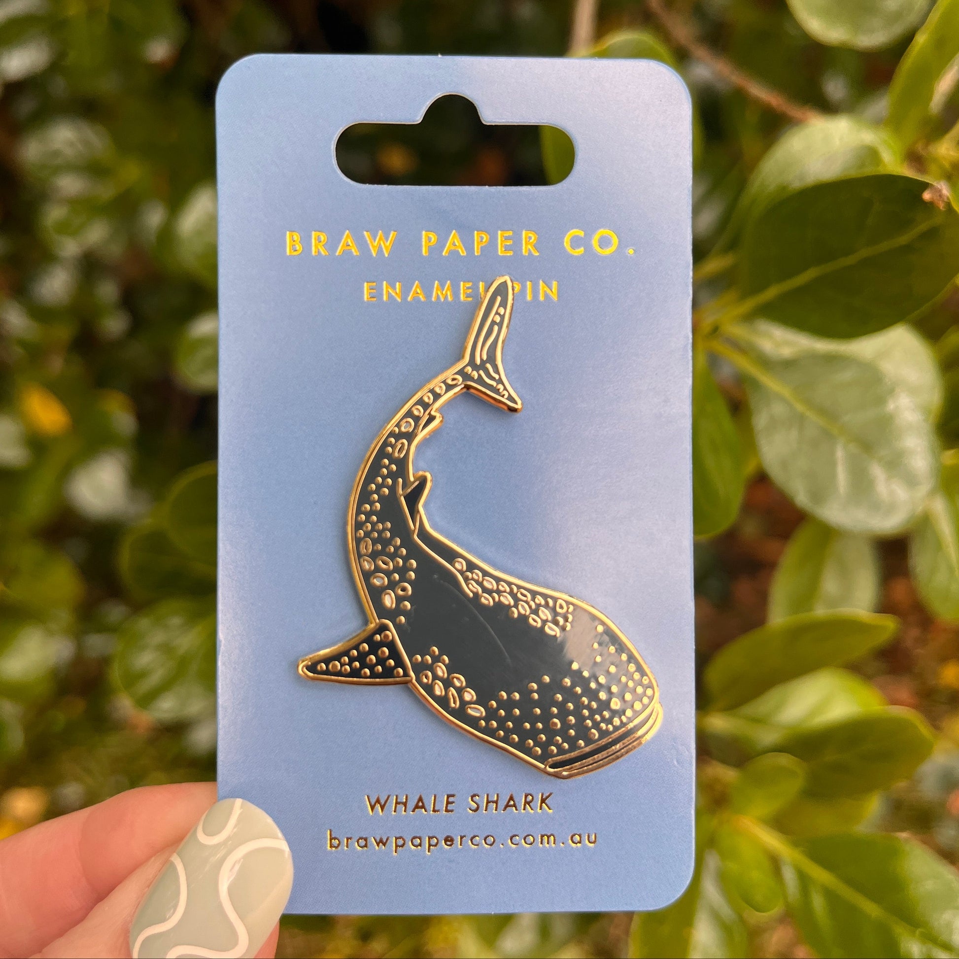 Gold whale shark enamel pin on a blue card with 'Braw Paper Co.' branding, held against a green leafy background.