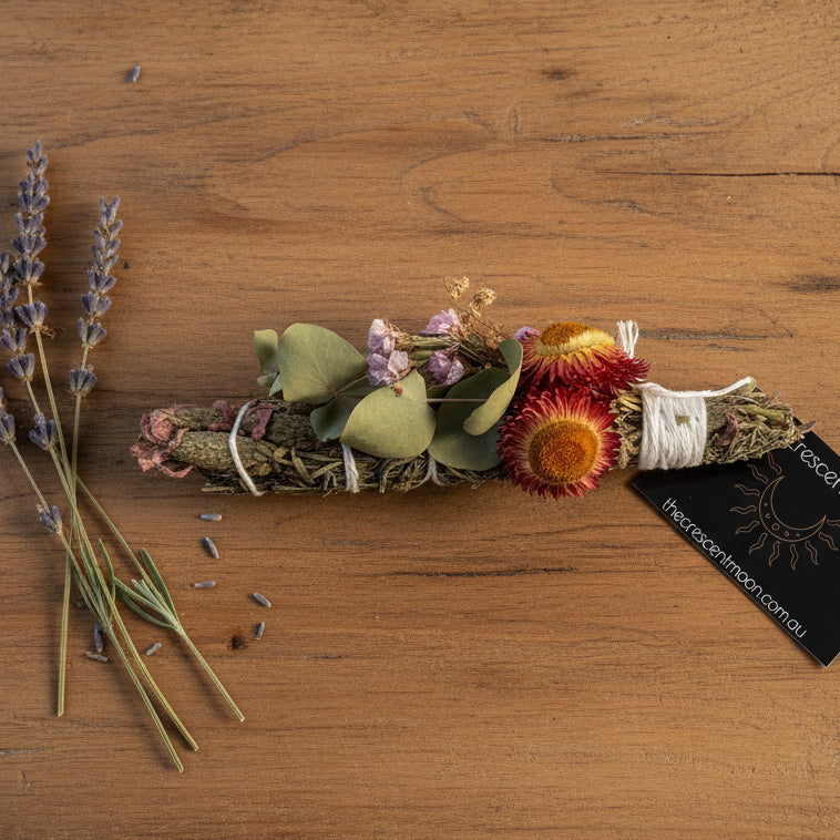 A dried flower and herb smudge bundle with green leaves, pink and red flowers, and white wrapping, shown upright on a white background with a black brand tag reading The Crescent Moon.