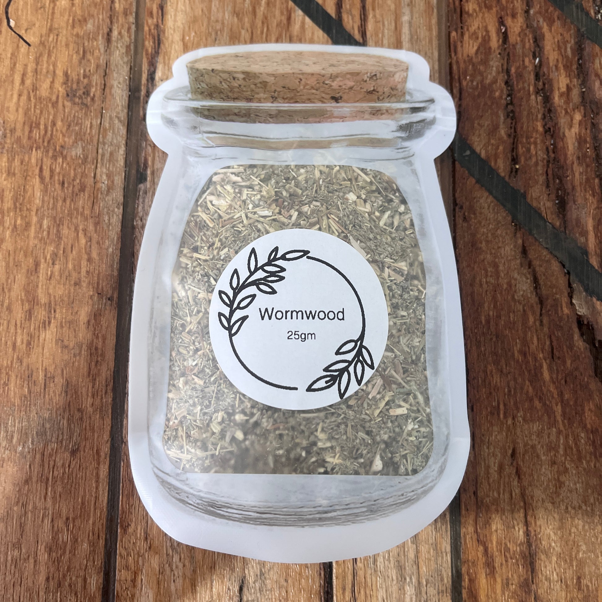 A small clear packet and a round white label reading 'Wormwood 25gm', filled with dried wormwood herb, placed on a wooden surface.