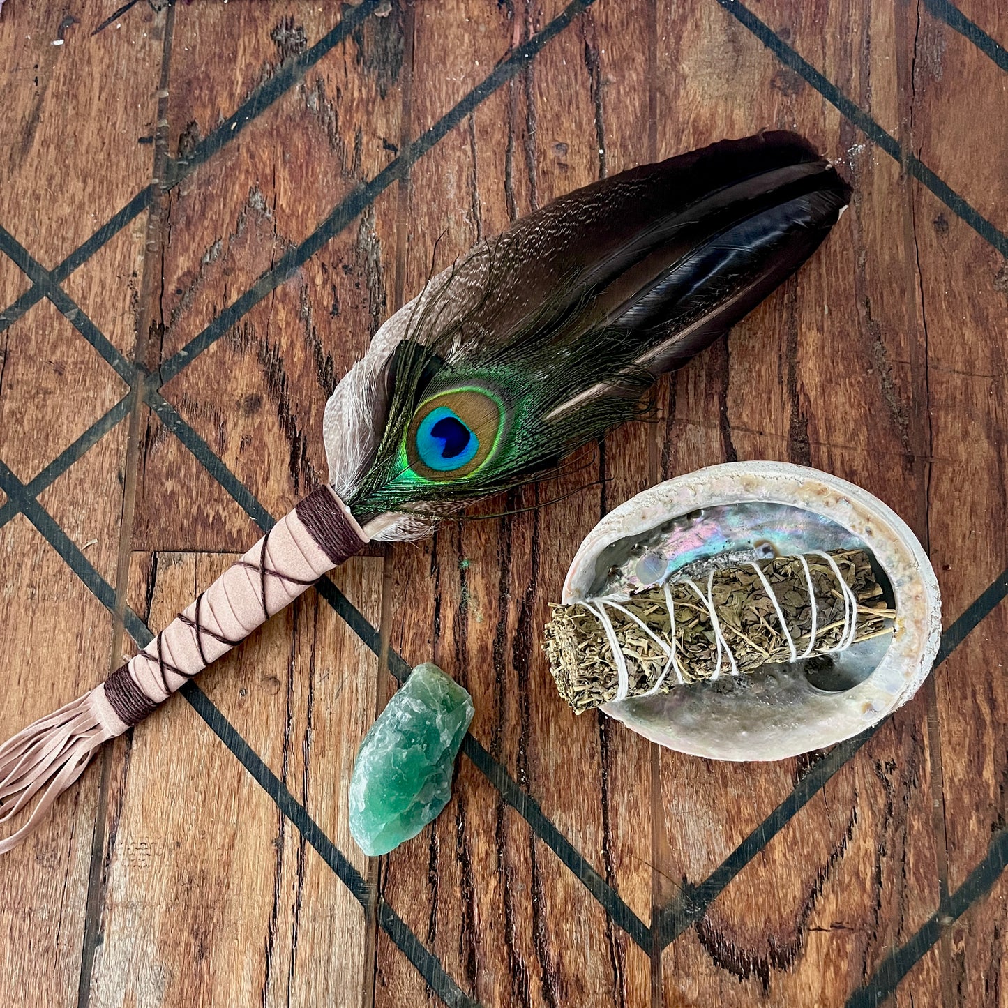 A styled flat-lay of a handmade smudge stick holder featuring a dark brown peacock feather, a sage bundle wrapped in white string inside a white shell dish, a green crystal, and a wrapped bundle on a dark wood background.