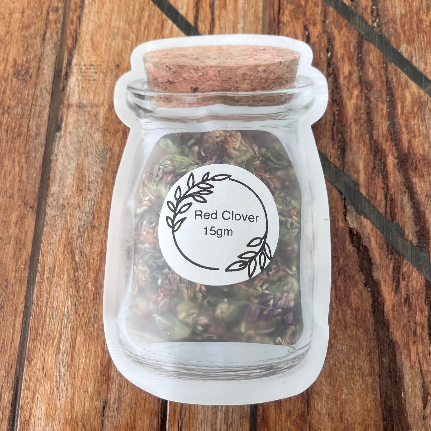 A small clear packet with a white circular label reading 'Red Clover 15gm,' filled with dried red clover, placed on a wooden surface.