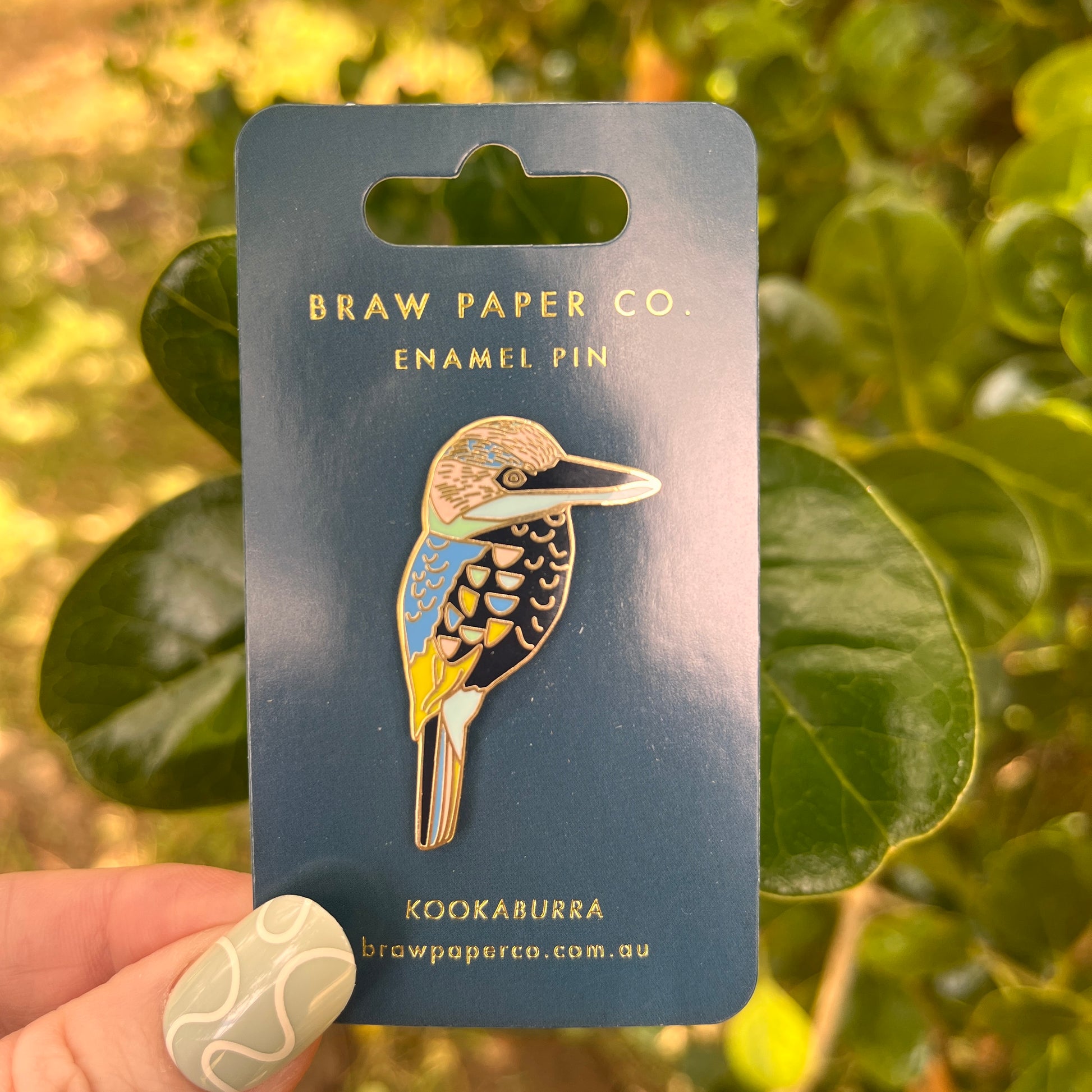 Enamel pin of a kookaburra held on a card by Braw Paper Co. with green leaves in the background.