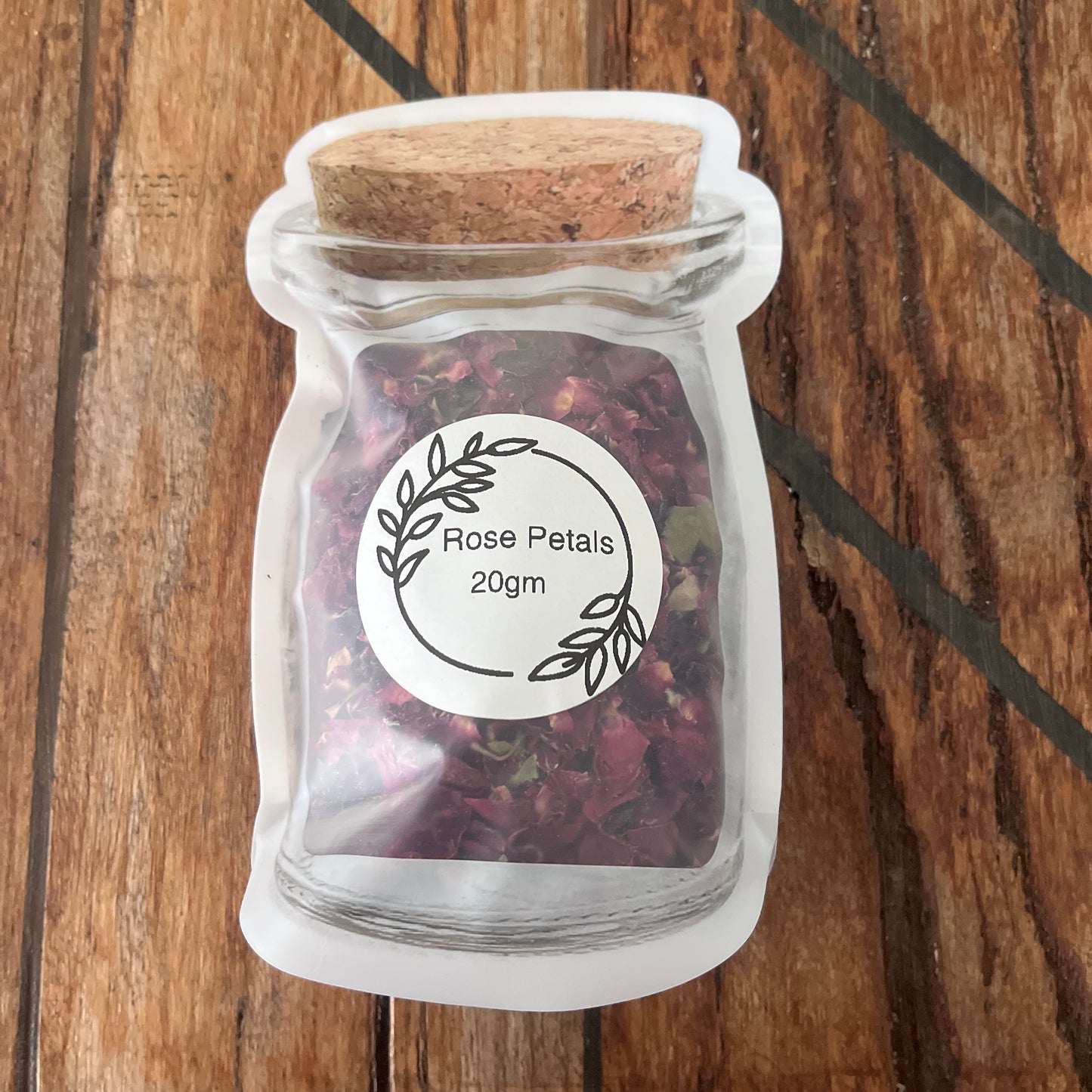 A small clear reusable plastic packet  filled with dried rose petals, featuring a round white label that reads 'Rose Petals 20gm', placed on a wooden surface.