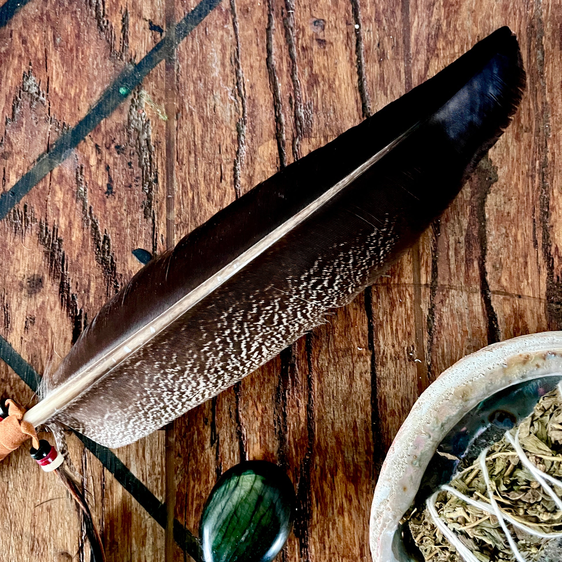 A dark brown and black smudging feather with a white center stripe lies diagonally on a rustic wooden surface, alongside a small stone and a bowl of dried herbs.