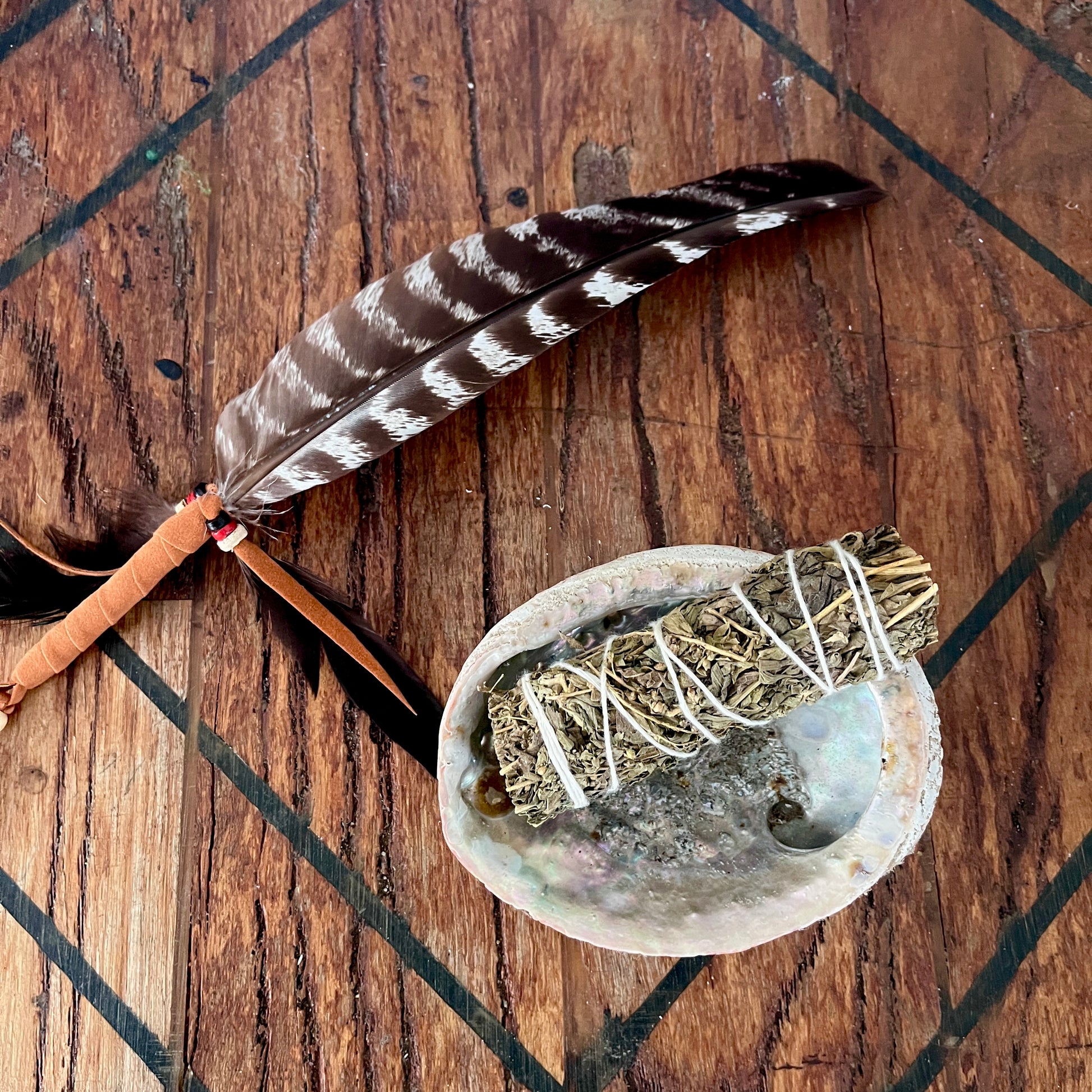 A natural brown and white patterned feather with a tan wrapped handle lies beside a sage smudge bundle wrapped in white string, placed on a polished abalone shell over a dark wood surface.