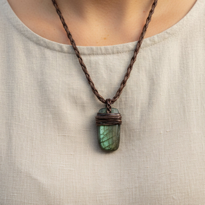 A handmade pendant necklace featuring a polished labradorite stone wrapped in dark macrame thread, hanging from a braided dark brown cord