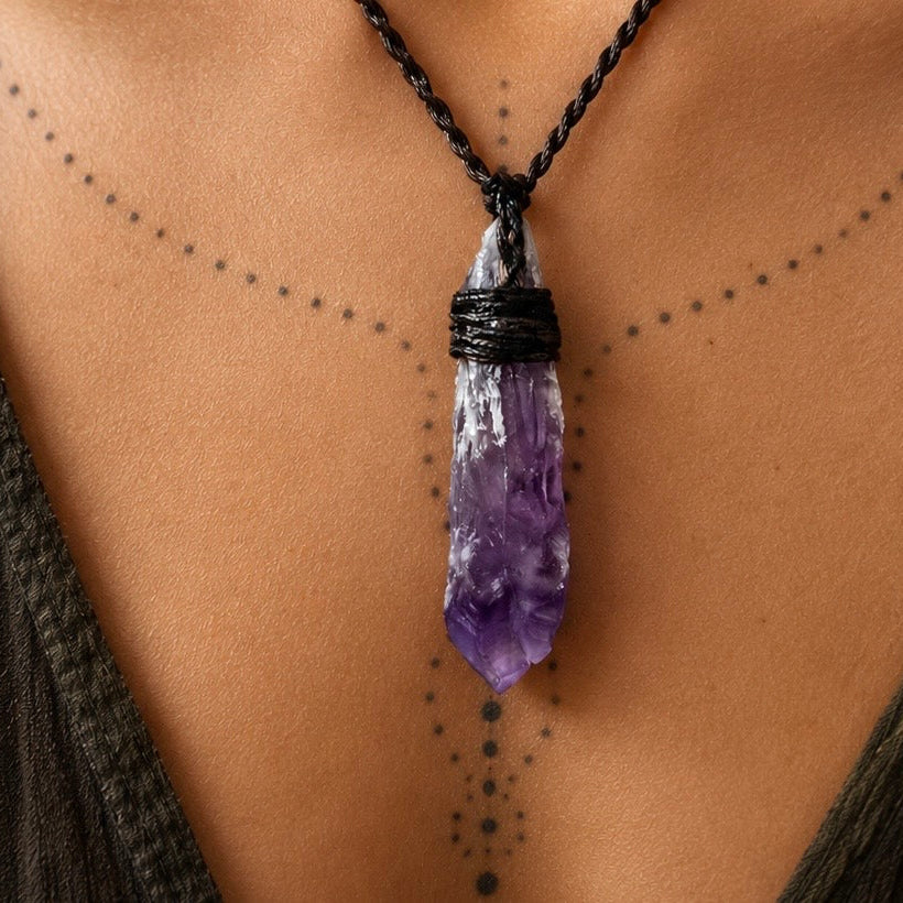 A close-up of a person's chest wearing a black cord necklace with a purple amethyst crystal point pendant. The person is wearing a dark brown draped top and has black geometric tattoo dots on their skin.