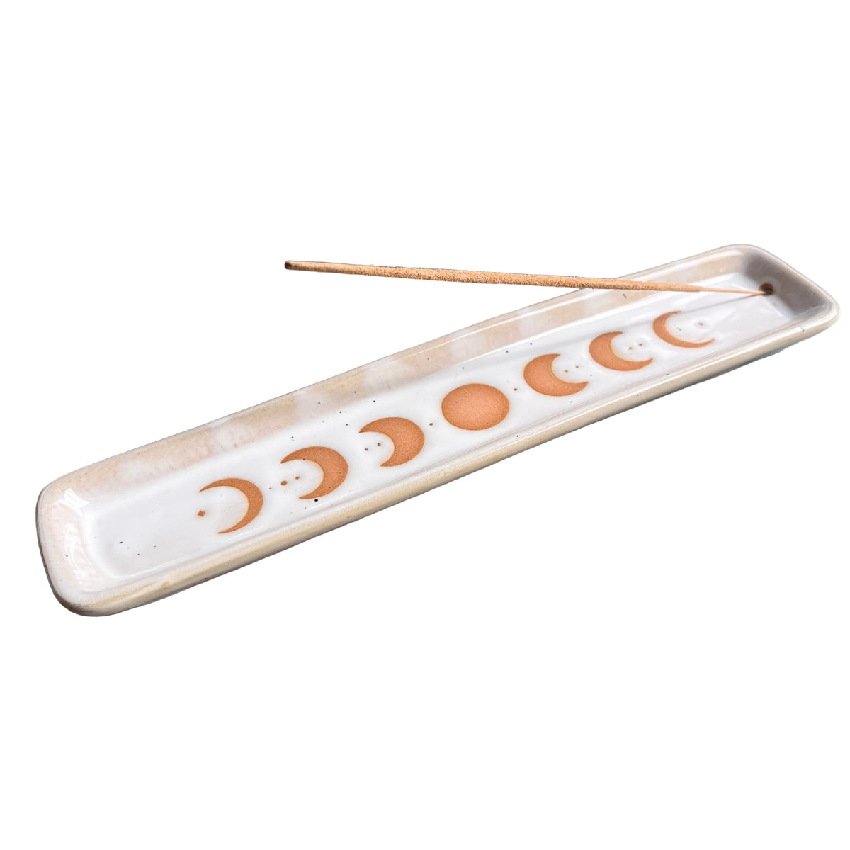 A rectangular ceramic incense holder with a moon phase design, containing a single incense stick, resting on a dark textured surface.