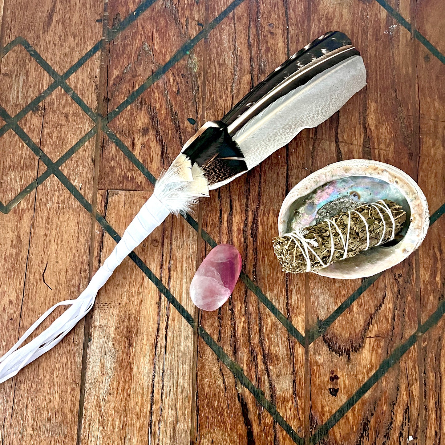 A handcrafted feather wand with a black and white patterned feather and white string tassels, displayed on a rustic wooden surface alongside a small pink crystal, a white shell containing a sage bundle wrapped in string, and a textured wooden tray.