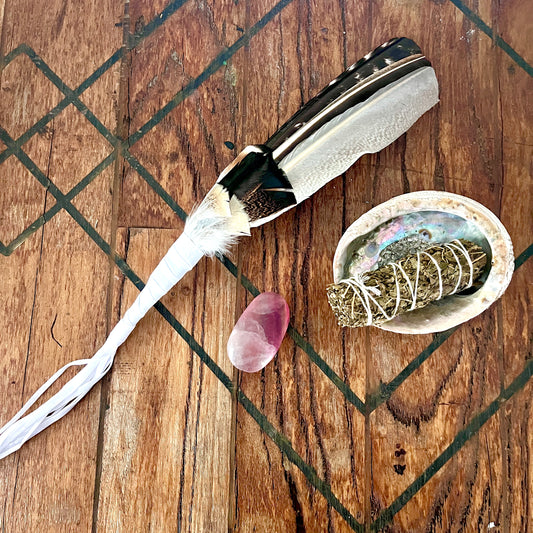 A handcrafted feather wand with a black and white patterned feather and white string tassels, displayed on a rustic wooden surface alongside a small pink crystal, a white shell containing a sage bundle wrapped in string, and a textured wooden tray.