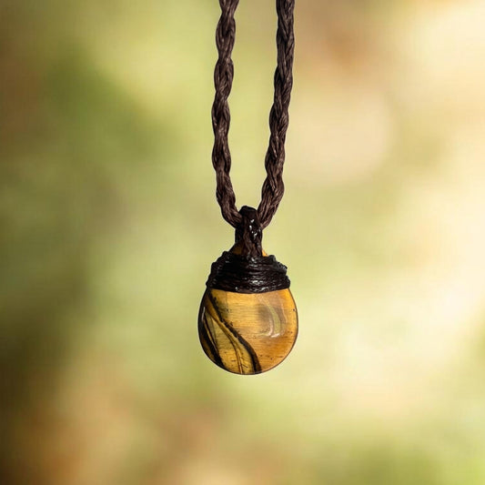 A close-up of a pendant necklace featuring a polished tiger's eye bead with dark brown wrapping at the top, suspended from a dark brown braided cord against a soft, blurred green and yellow bokeh background.