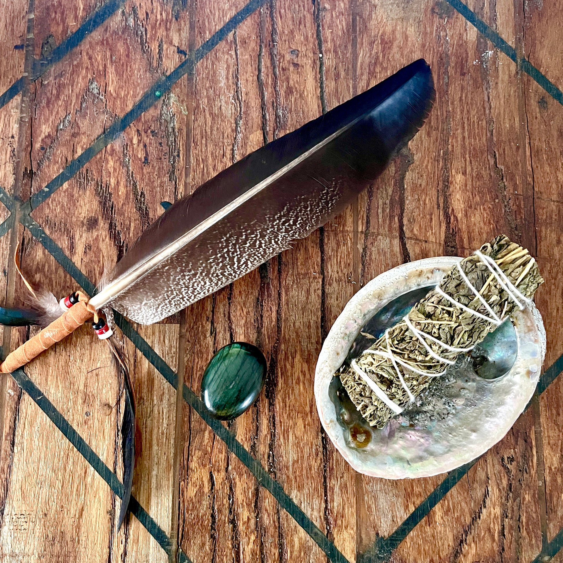 A dark brown feather with a small orange tip and bead detail lies on a rustic wooden surface. Next to it is a green polished stone and a white abalone shell holding a sage smudge bundle wrapped in white string.