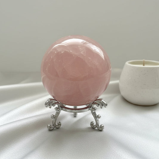 Pink crystal ball on a silver stand next to a candle on a light surface.