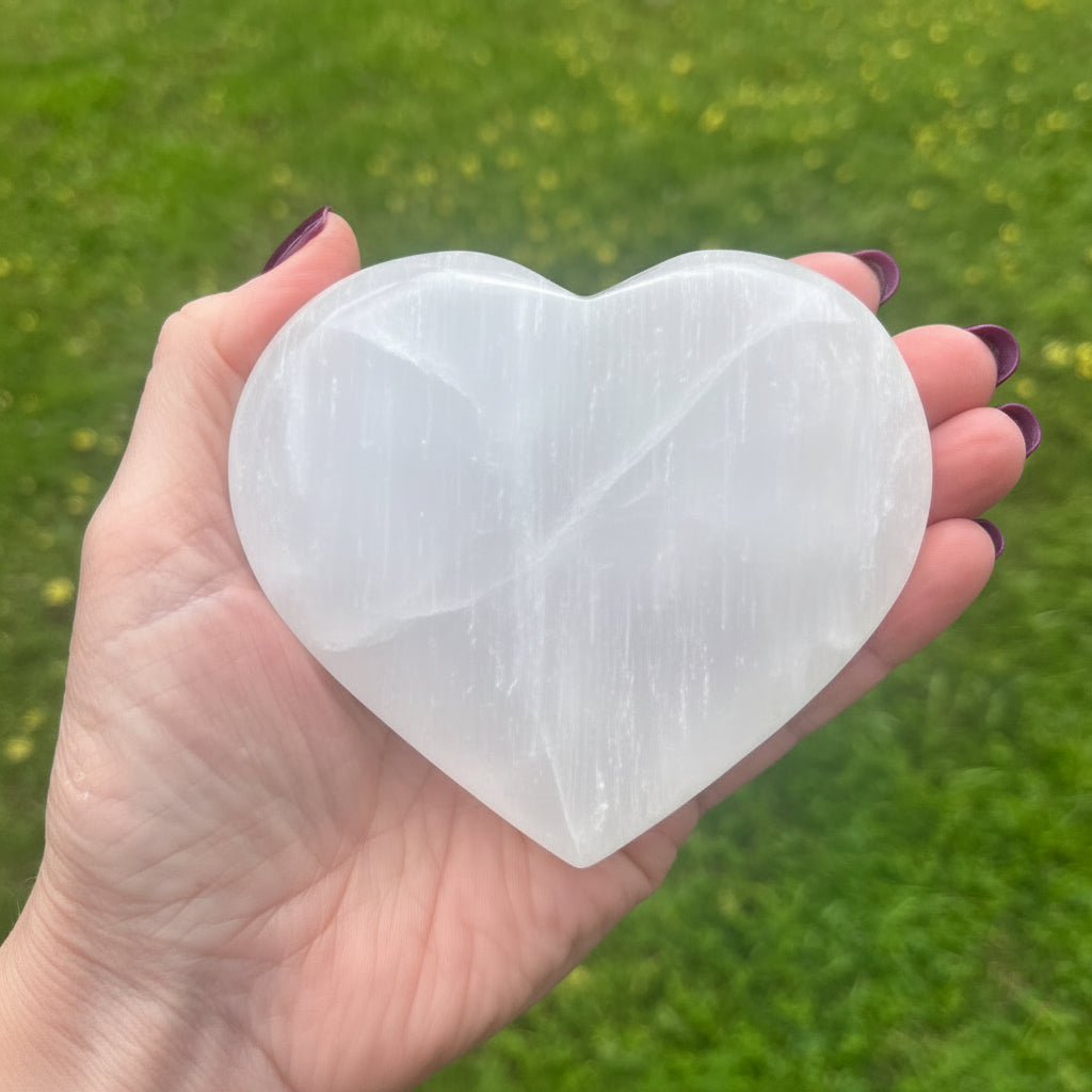 Heart-shaped selenite crystal held in a hand with a grassy background