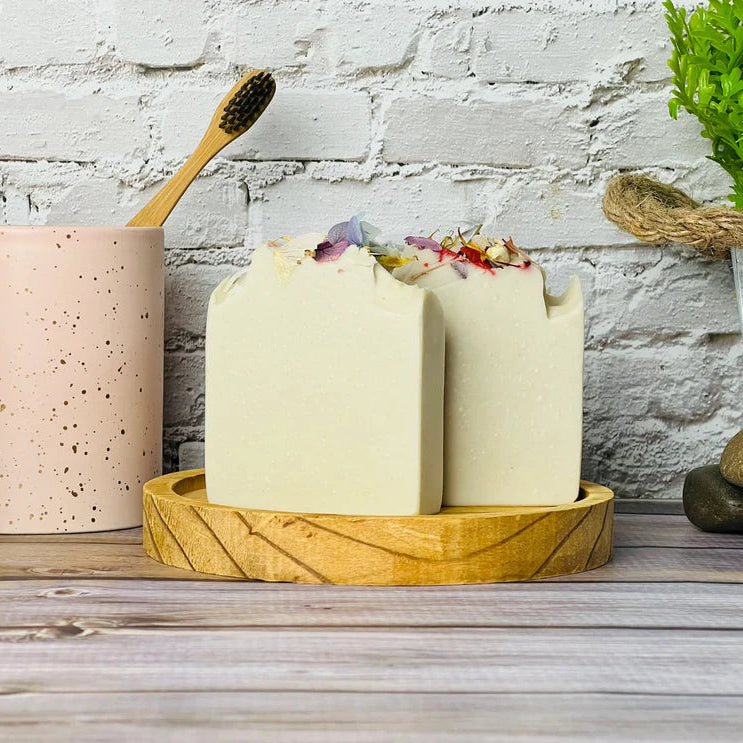 Two bars of soap on a wooden stand with a pink container and toothbrushes against a stone wall.