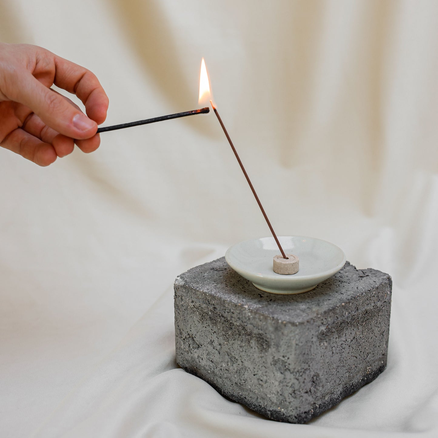 A hand holds a lit incense stick over a small white dish resting on a textured gray stone block, set against a soft cream fabric backdrop.