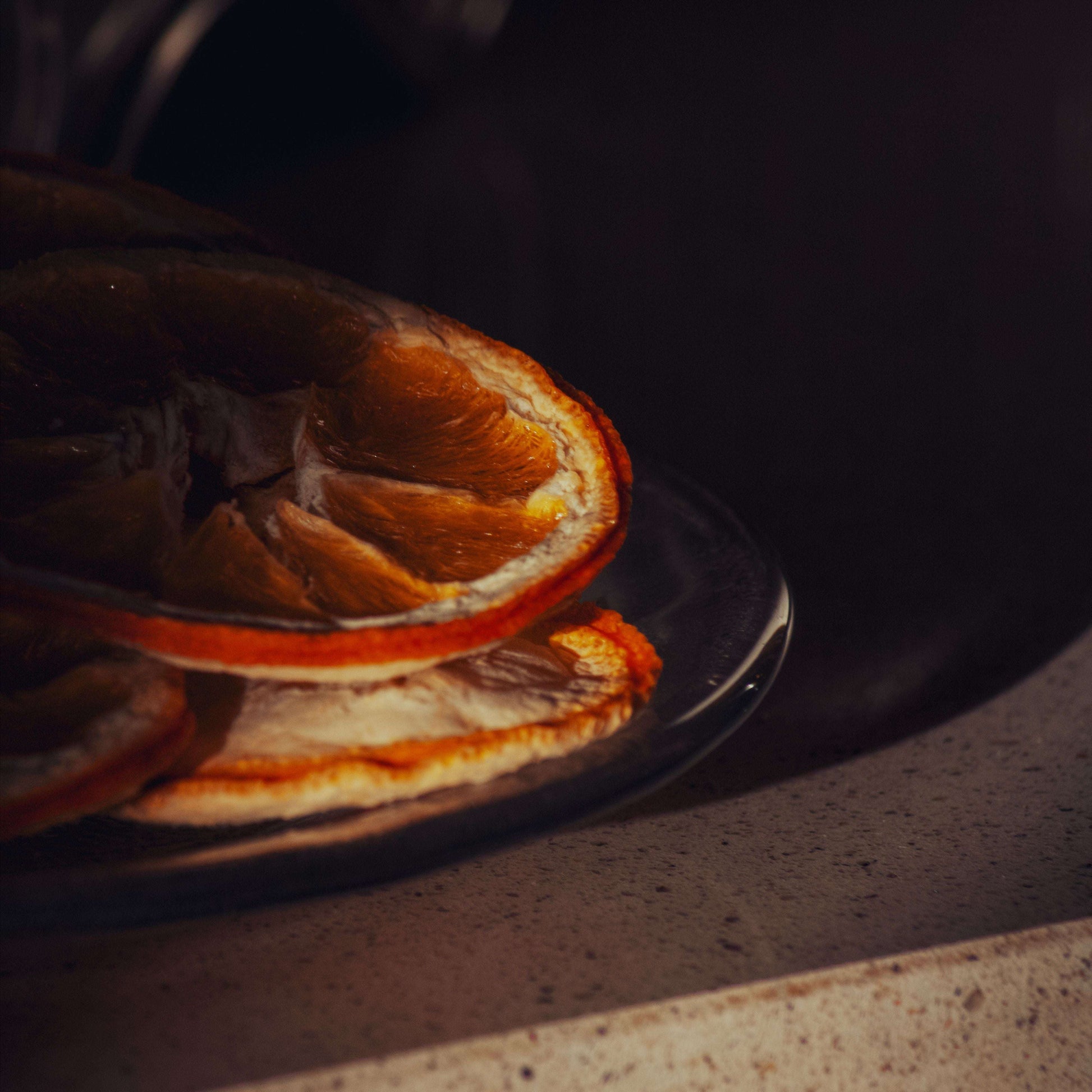 Dried orange slices stacked on a dark plate, set on a speckled beige countertop in a dimly lit kitchen setting.
