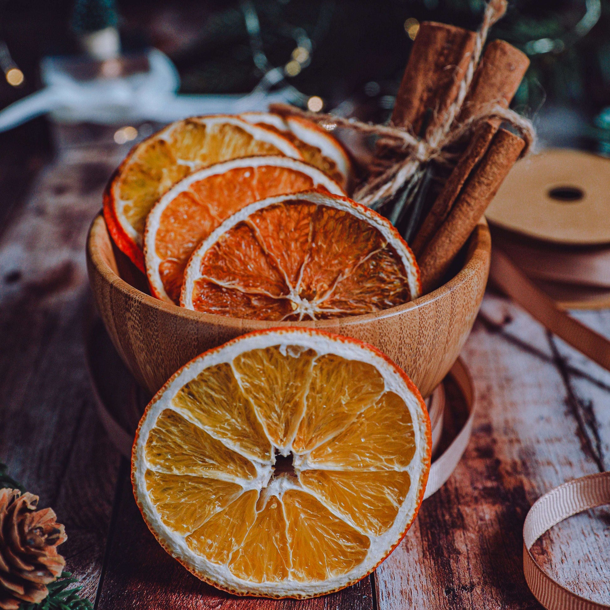 Dried citrus slices and cinnamon sticks arranged in a small wooden bowl on a rustic wooden surface, with a blurred holiday scene in the background.