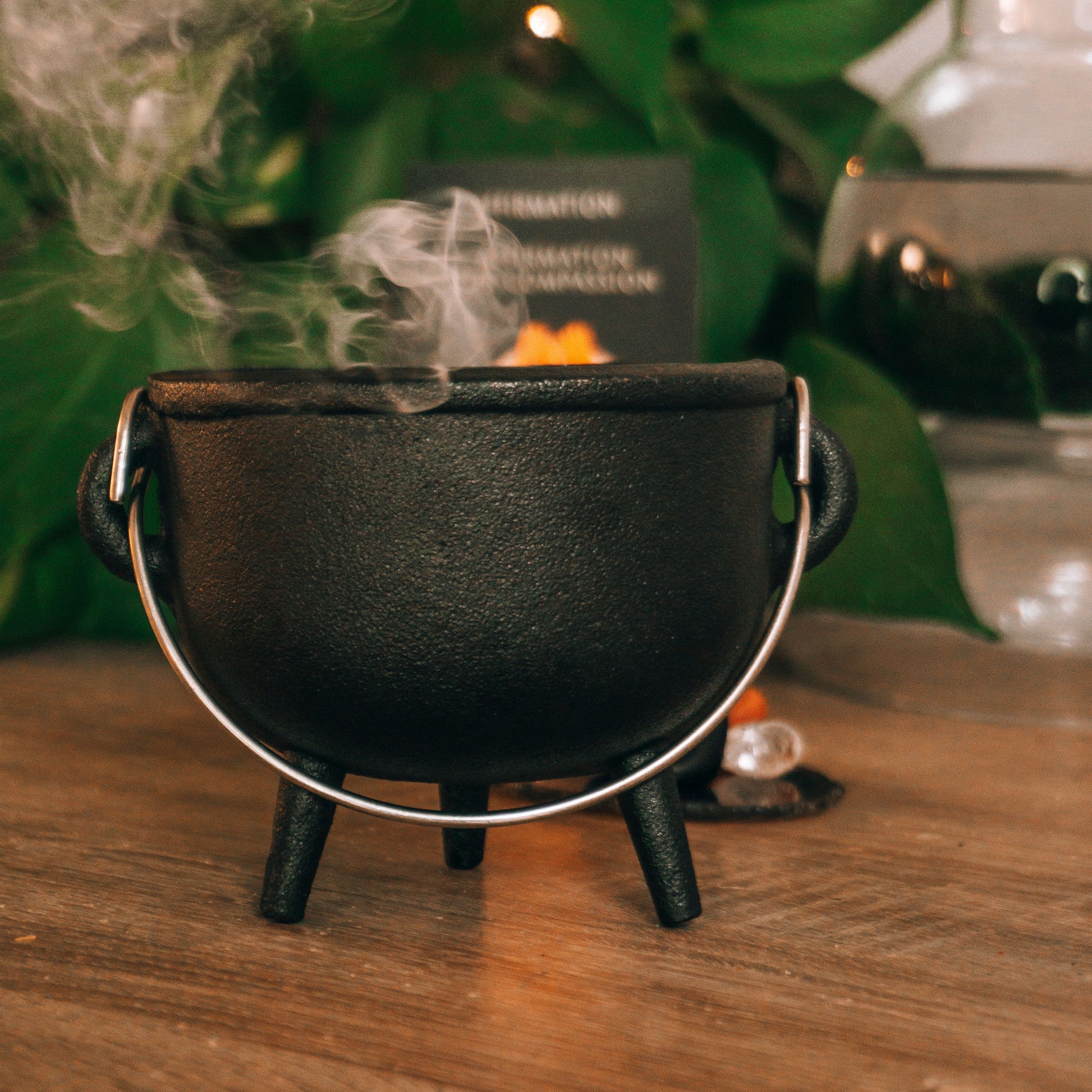 A miniature black cast iron cauldron with a silver metal handle sits on a wooden table, with white steam rising from inside. The background features green houseplants and warm, soft-focus lighting.