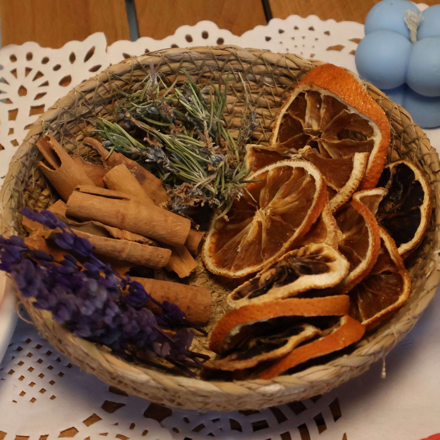 A woven basket filled with dried botanicals including orange slices, cinnamon sticks, lavender, and rosemary, arranged on a white lace doily over a wooden table.