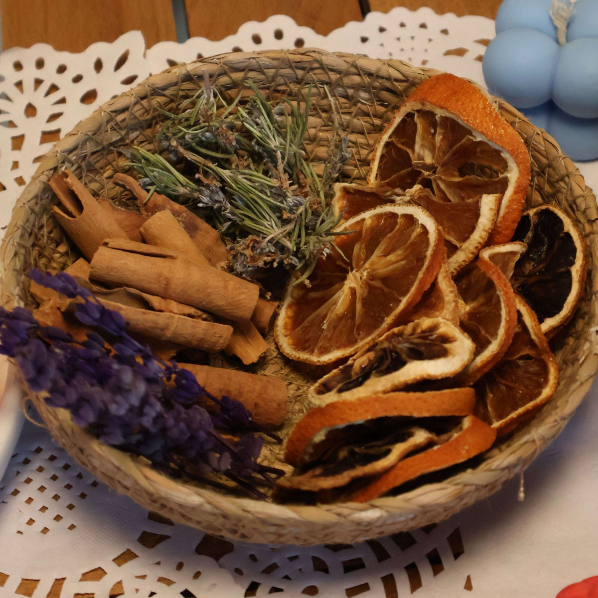 A woven basket filled with dried botanicals including orange slices, cinnamon sticks, lavender, and rosemary, arranged on a white lace doily over a wooden table.