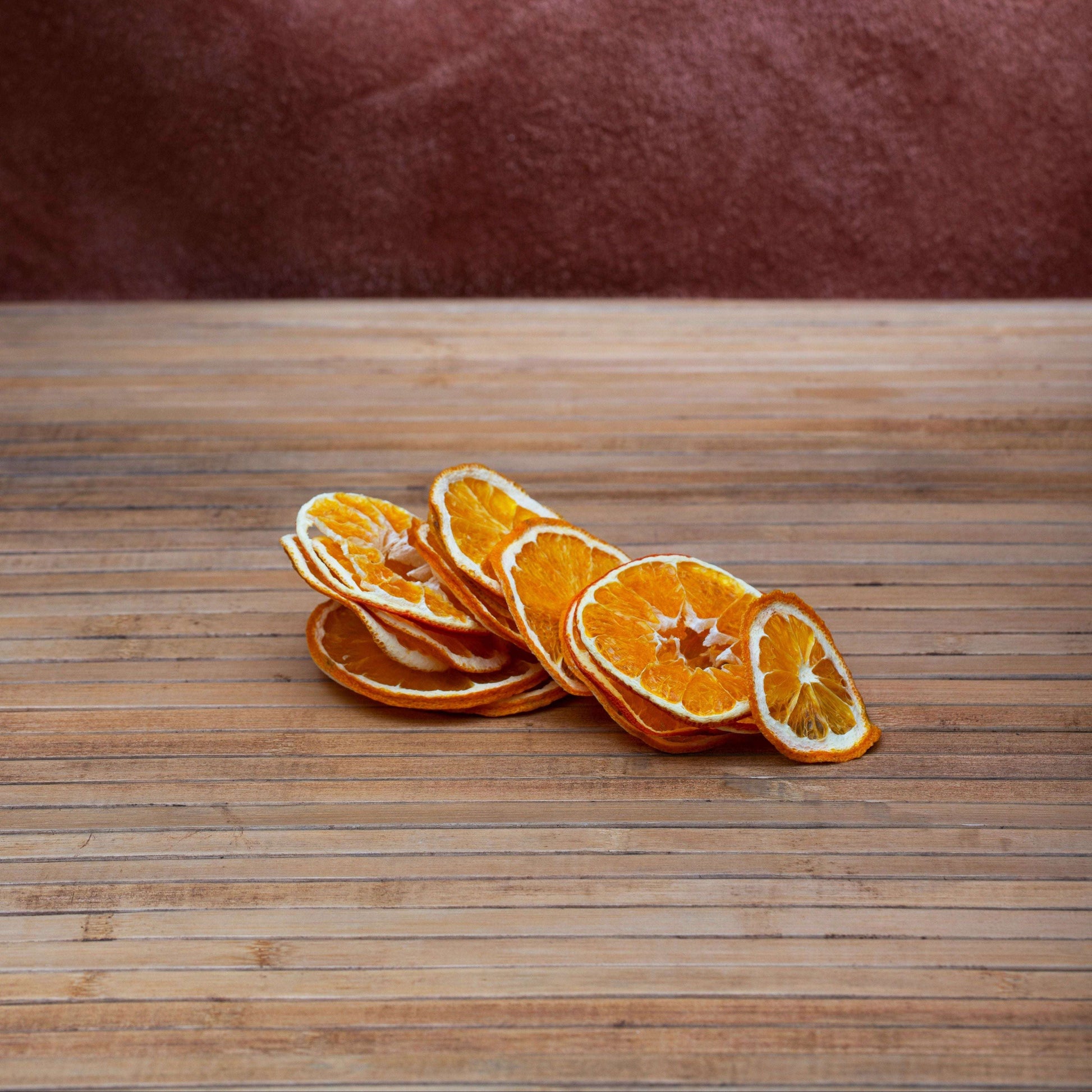 A small pile of dried orange slices with visible white pith, arranged on a wooden tabletop against a mottled reddish-brown background.